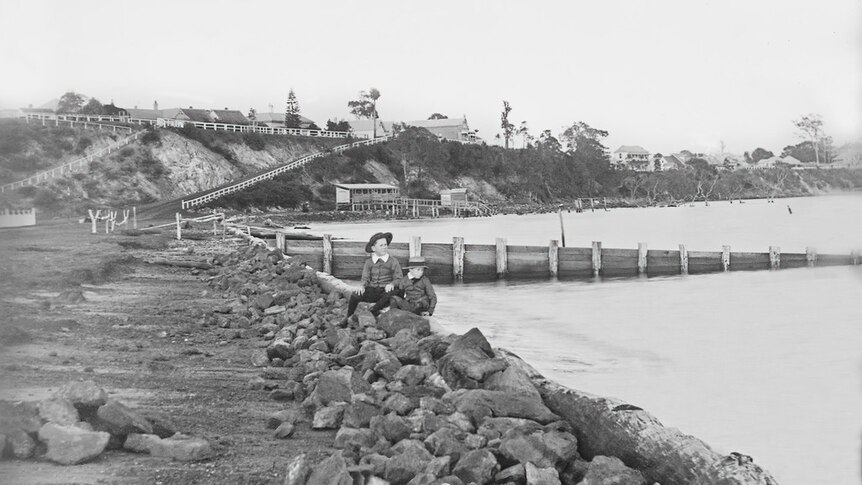 A black and white photograph of a rocky shoreline. A boy sits on the rocks.