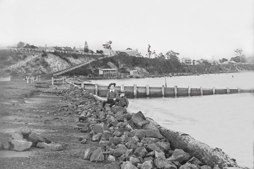 A black and white photograph of a rocky shoreline. A boy sits on the rocks.