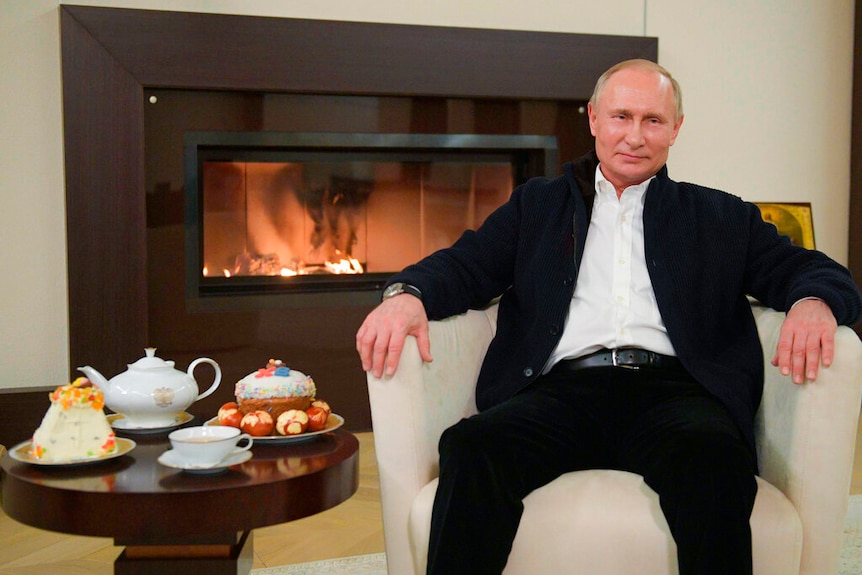 Vladimir Putin sits in front of a fire with a pot of tea, cake and easter eggs beside him.