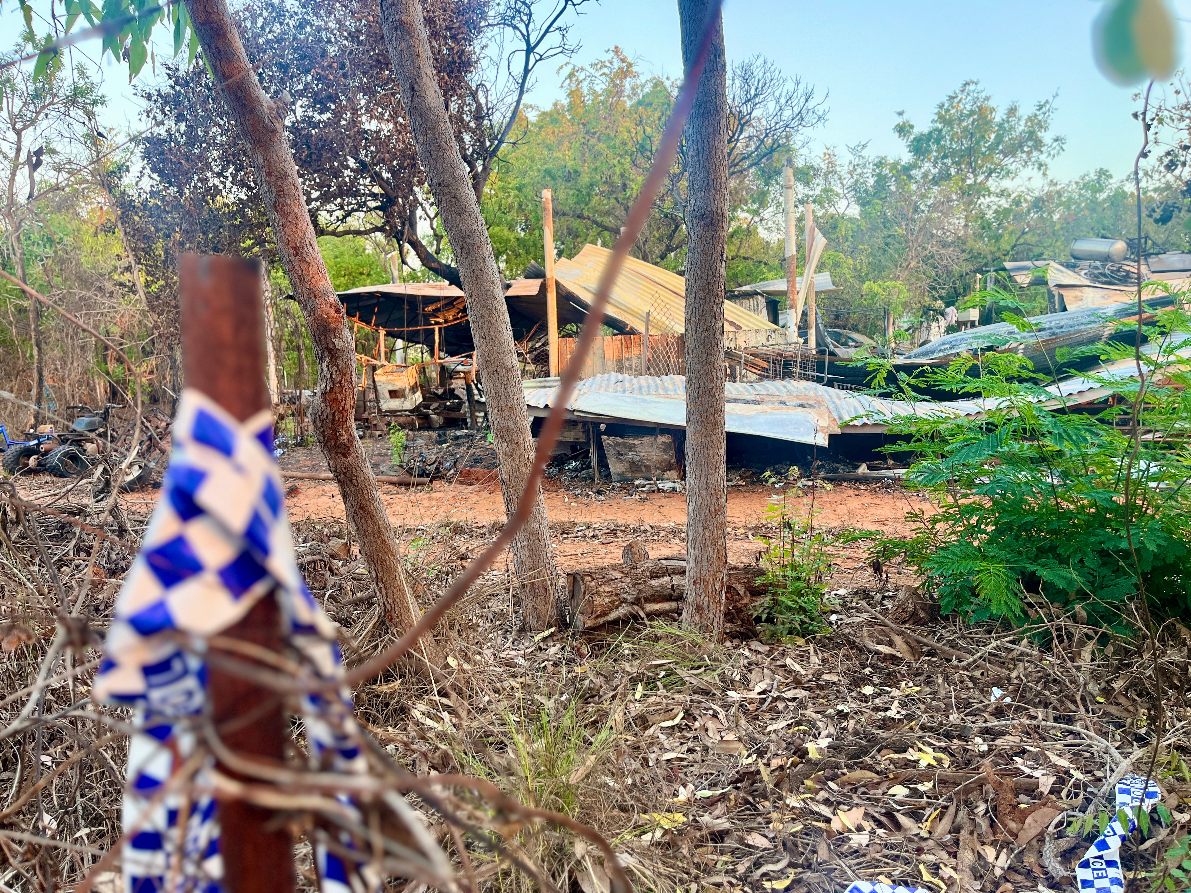 Police tape around a fence in front of a burnt shed and wreckage of a caravan.