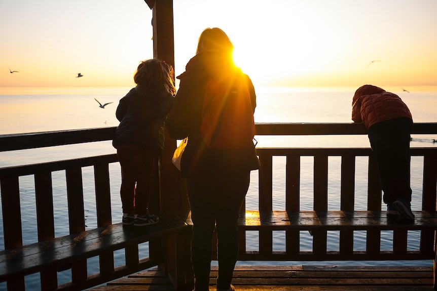 A mother and children silhouetted on a balcony looking out to see as the sun sets.