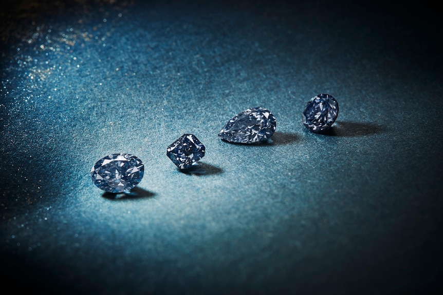 A stunning array of four blue, radiant-cut diamonds — an oval, an oblong, a teardrop and a round — spotlit on a blue cloth  