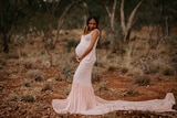 A pregnant Indigenous woman poses with her arms around her bell, wearing a pink gown with lace detail on red rocky landscape
