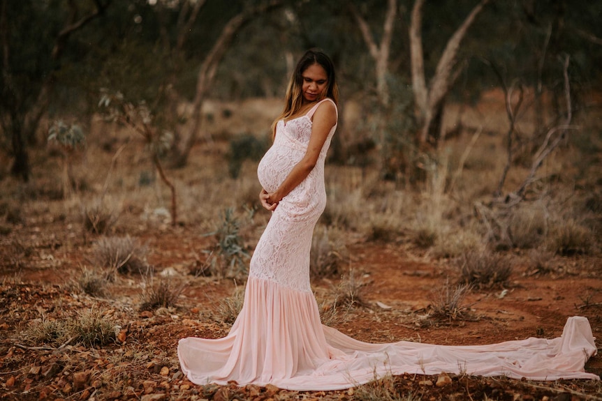 A pregnant Indigenous woman poses with her arms around her bell, wearing a pink gown with lace detail on red rocky landscape