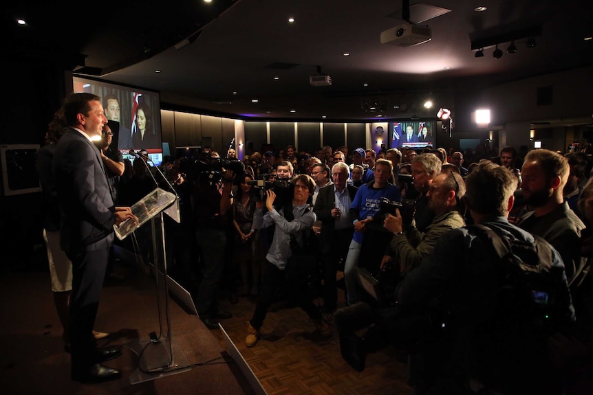 Matthew Guy delivers a speech as a large group from the media film and photograph him.