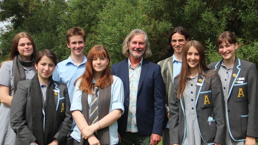 Mid-shot of a man with long hair, wearing a blue jacket, surrounded by young people in school uniform, trees in the background.