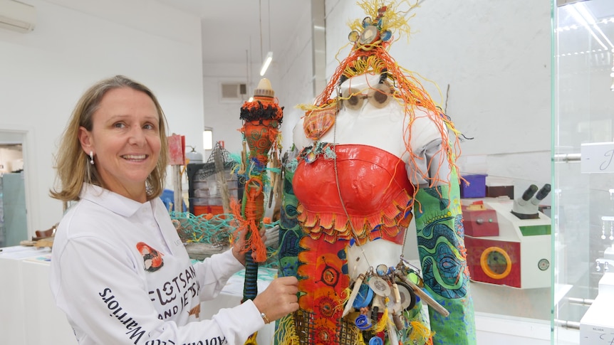 smiling woman wearing a white top standing next to costume of red bodice and pieces of jewellery