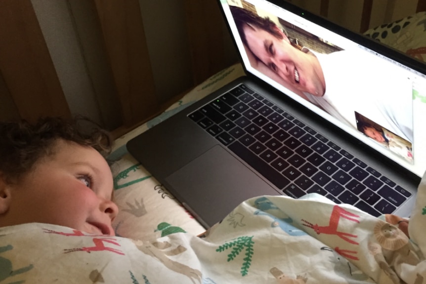 A toddler lays in bedding looking at a laptop screen with a side-on image of a smiling man.