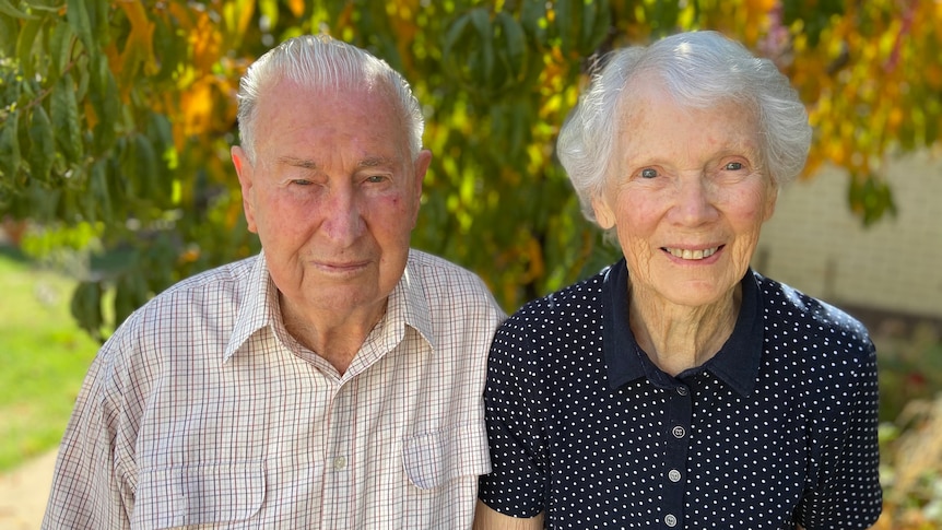An older man and woman, with silver hair and wearing button-down shirts, with green and yellow leaves in background.