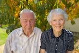 An older man and woman, with silver hair and wearing button-down shirts, with green and yellow leaves in background.