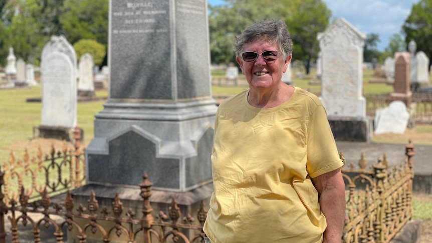 A lady in a yellow shirt and glasses smiles in front of a tombstone.