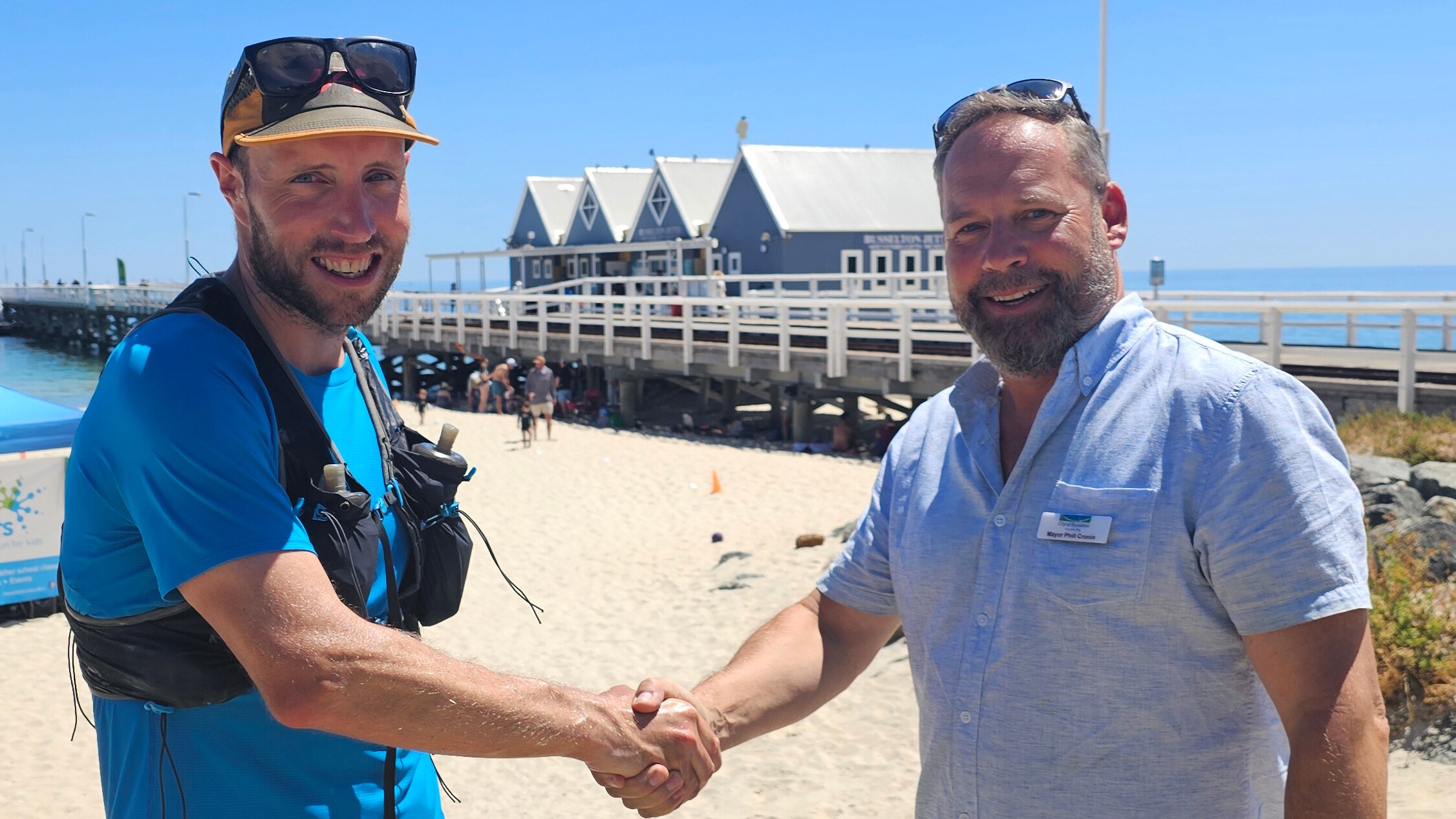 Two men shake hands in front of a timber ocean jetty. 