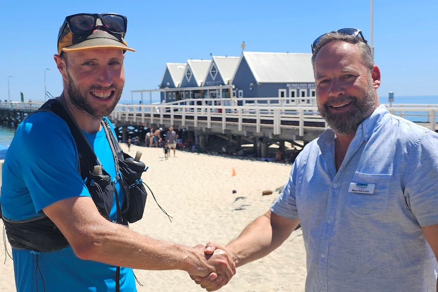 Two men shake hands in front of a timber ocean jetty. 