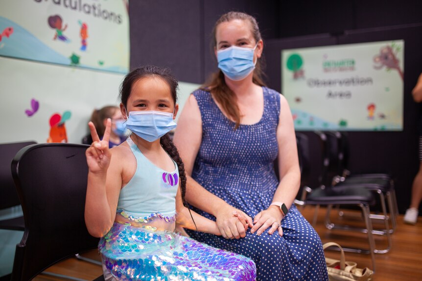 A child holds up two fingers and smiles behind a mask, sitting with their parent in a bright vaccination hub room.
