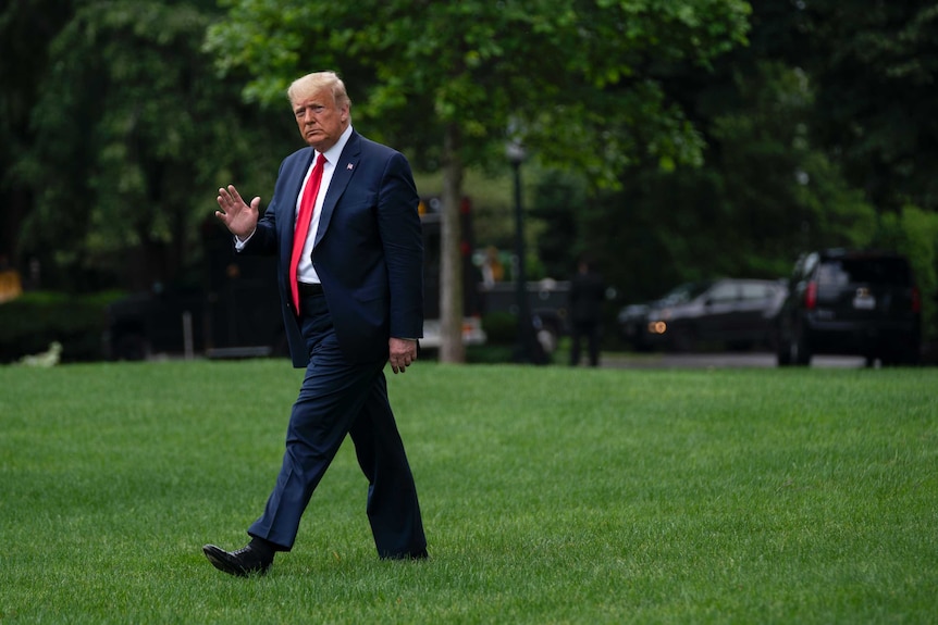 A man in a dark suit and red tie waves with his right hand as he walks across a verdant, well-kept lawn.