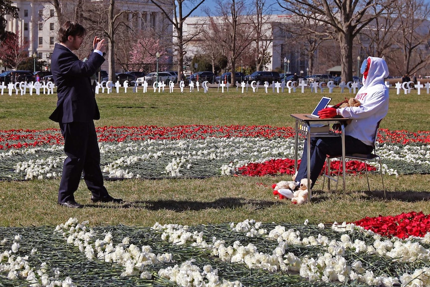 A man stands on a lawn with a camera photographing a dummy with a mirror for a face.