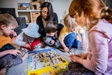 Children and staff are seen around a rug while playing at a childcare centre.