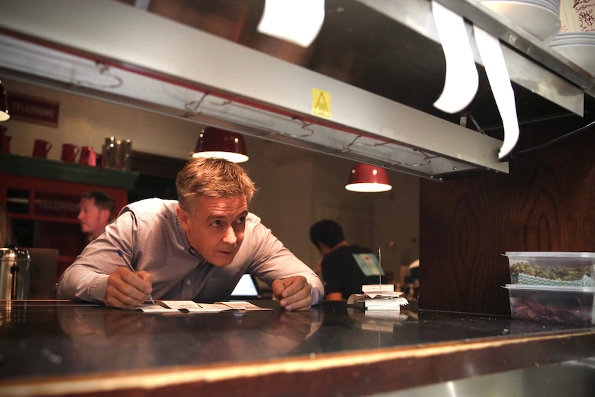 Martin James leans across a counter with a pen in his hand inside his restaurant in Queenstown