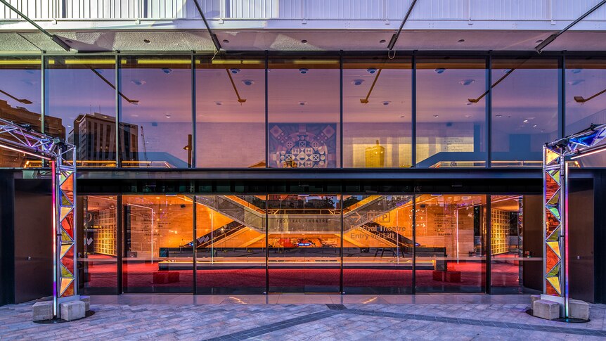 The glass wall to the foyer of a theatre behind it with Indigenous themed artwork