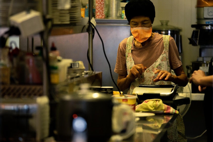 A woman in a mask and apron in a small commercial kitchen.