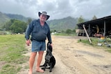 A man in a dark coloured workshirt, denim shorts and no shoes patting a dog on a rural property.