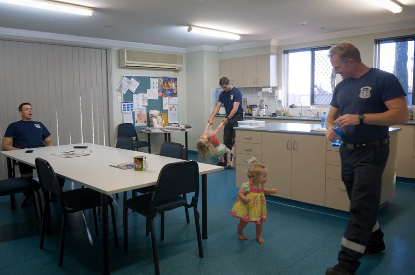 A younger toddler wanders through and another young girl swings off the arms of a fireman inside a workplace breakroom.