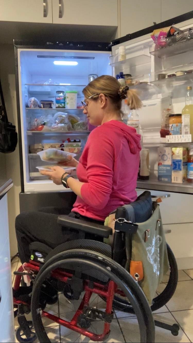 A woman in a pink hoodie looks through the fridge while sitting in a wheelchair.