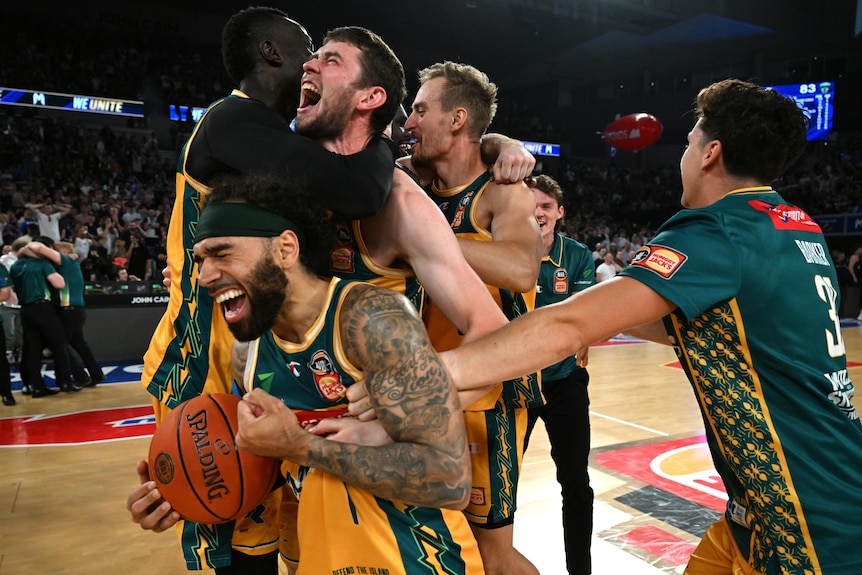 A group of Tasmania NBL players cheer, cry and embrace on the court after winning the title. 