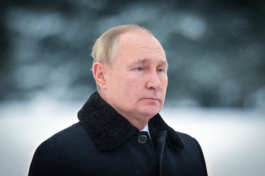 Vladimir Putin looking sombre, dressed in a black coat while standing in the snow 