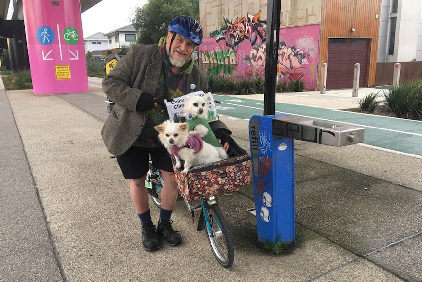 An older man holds a bike with two small dogs in front basket, poster says chihuahuas for climate action