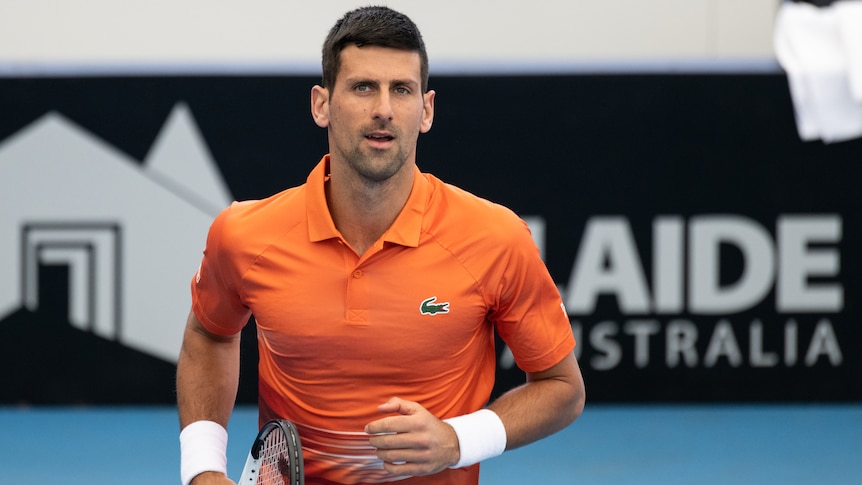 Tennis star Novak Djokovic looks at the camera as he walks on a tennis court carrying a racquet by his side.