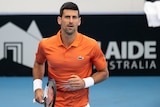 Tennis star Novak Djokovic looks at the camera as he walks on a tennis court carrying a racquet by his side.