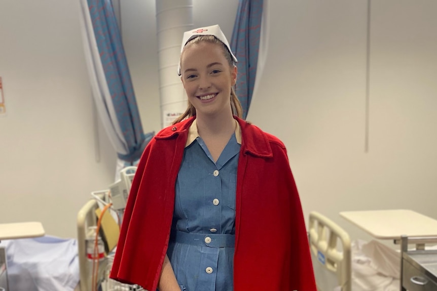 Audrey Sproules stands in a hospital ward dressed in a vintage blue nurses uniform with red cape.