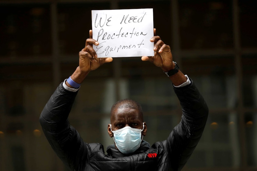 A man in a face mask holding up a sign reading "We Need Protection Equipment"