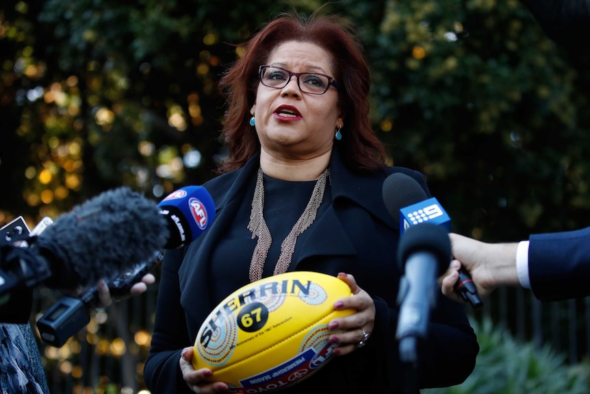 Tanya Hosch stands before an array of media microphones. She's holding and AFL Indigenous round Sherrin football.