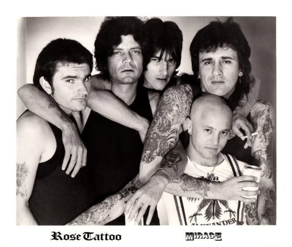A black and white photo of four dark haired men and a bald man standing with their tattooed arms around each others' shoulders.