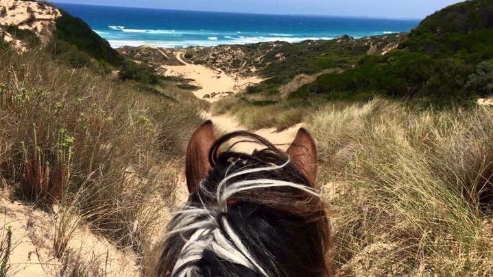 A woman rides a horse through a sandy track among scrub, with a rainbow visible overhead.