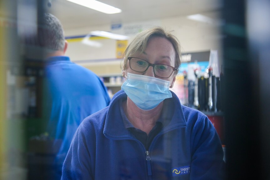 A woman is photographed through a store window. She is wearing a retail uniform and a facemask
