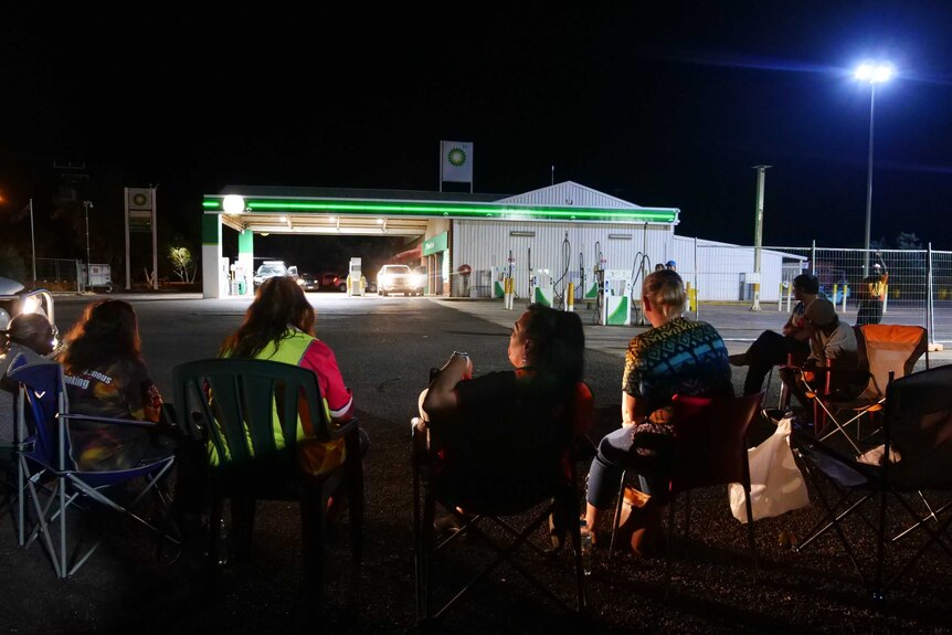 A group of women sitting on chairs with a service station in the background at night.