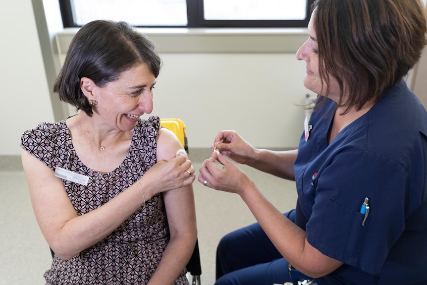 Gladys Berejiklian holds a cotton pad to her shoulder while a nurse gets ready to put a bandaid on it after a vaccine.
