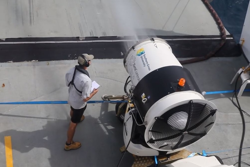 A high angle photo showing a man in ear muffs holding standing next to a large water canon on the back of a boat.