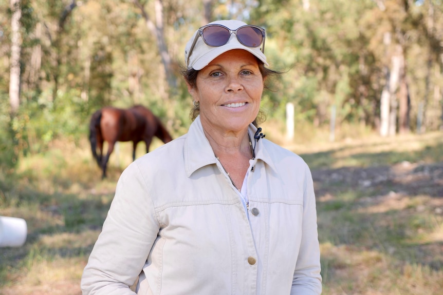 A woman stands in a paddock smiling with a horse in the background.