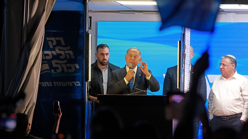 Benjamin Netanyahu speaking into microphone behind glass with three men standing around him