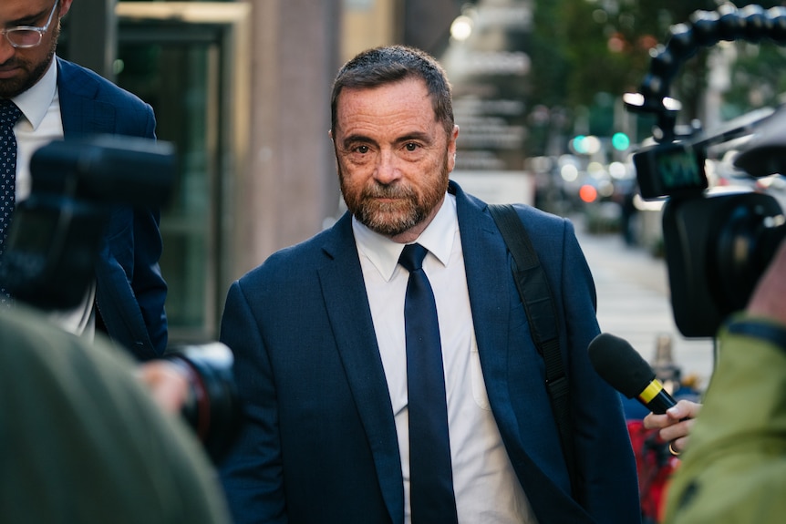 Michael wears a navy blue suit and tie with white shirt, walking down a city street with a media scrum surrounding him.