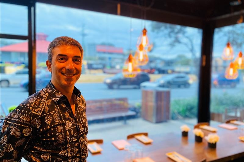 A smiling man wears a black and orange printed shirt, stands near a window in a restaurant with hanging lights, dining table.