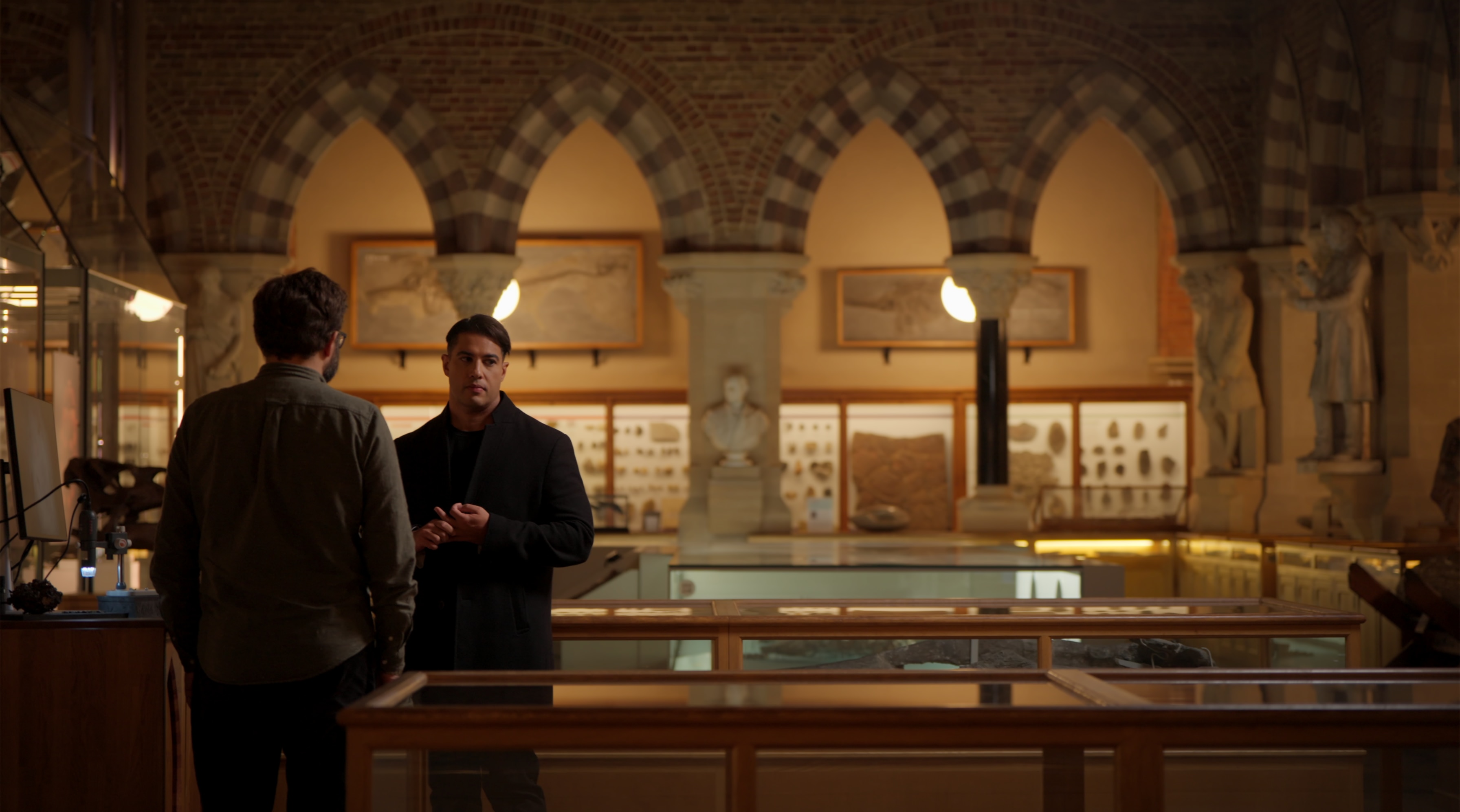 Marc stands in museum archives in dim lighting talking to an expert who has their back to us.