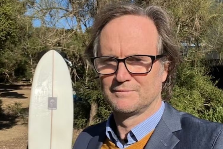A man wearing a suit stands outside in front of a surfboard. 