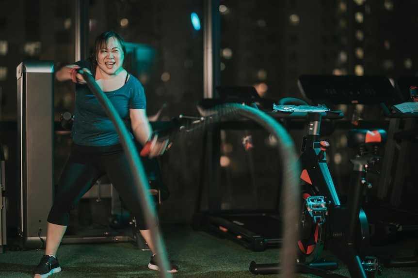 A woman grimaces while lifting long ropes alternately in each hand in a gym