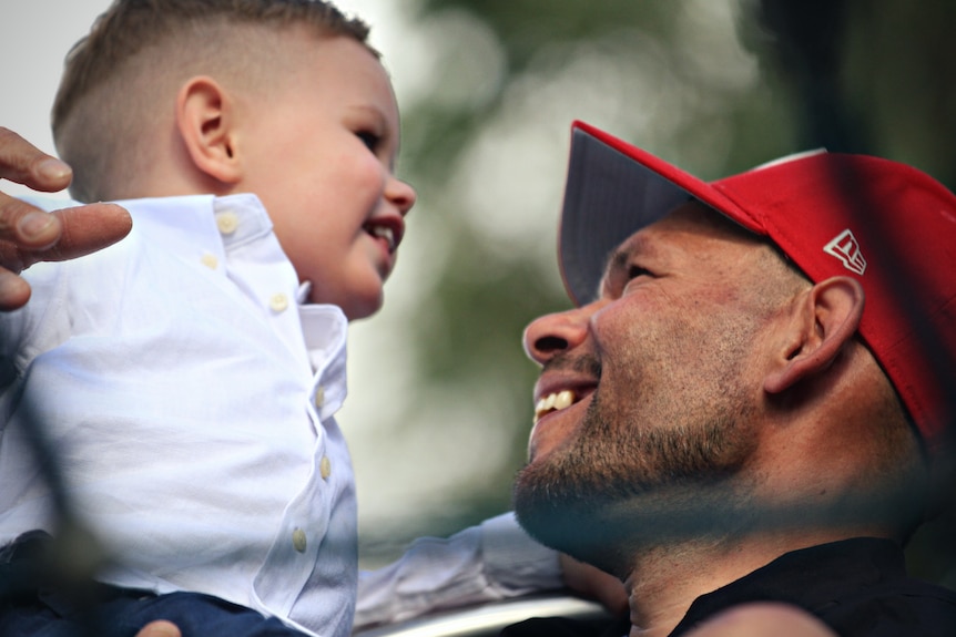 Man wearing a red cap holding up a young boy and smiling.