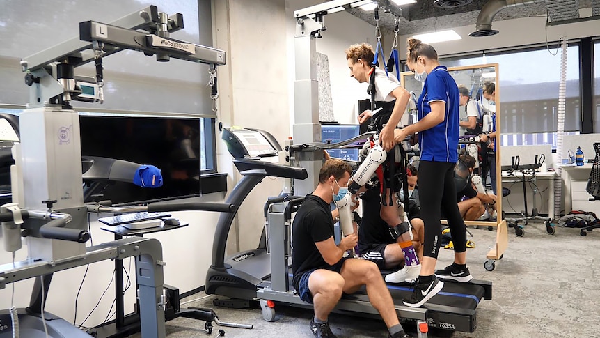 Kyle walks on the treadmill with the assistance of three physiotherapists while wearing the exoskeleton.